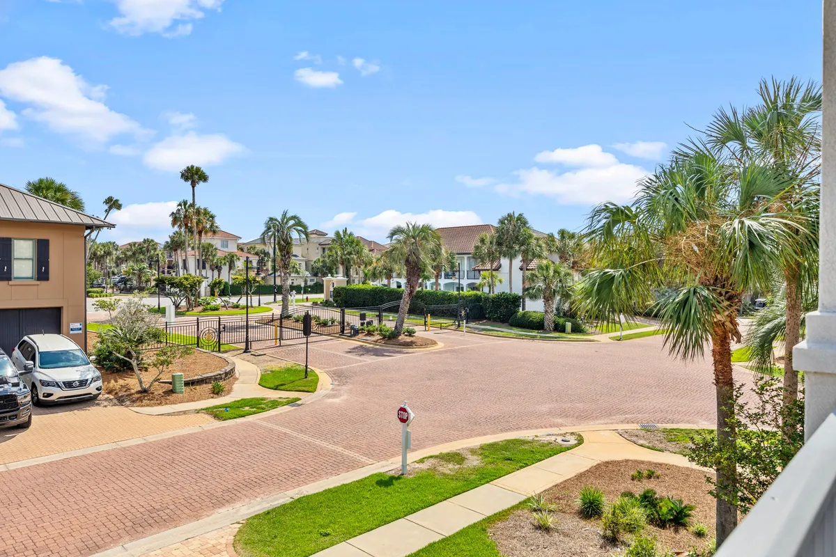 A relaxed neighborhood view with palm trees lining a paved road, a gated entrance, and residential buildings beyond. The setting suggests a safe, friendly community perfect for walks, bike rides, and socializing outdoors.