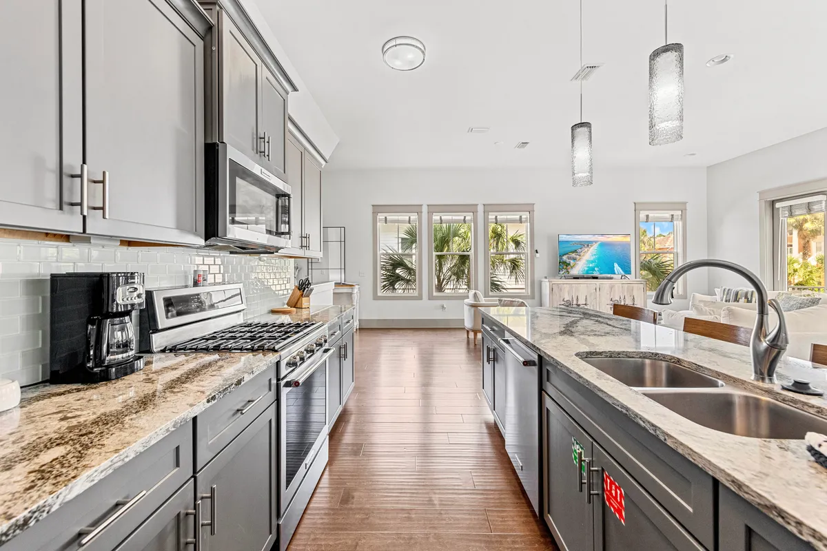This image showcases a modern kitchen with granite countertops, stainless steel appliances, a central kitchen island, and large windows that offer scenic views. The TV in the adjacent living area is also visible, highlighting the open-concept layout and the connected social spaces.