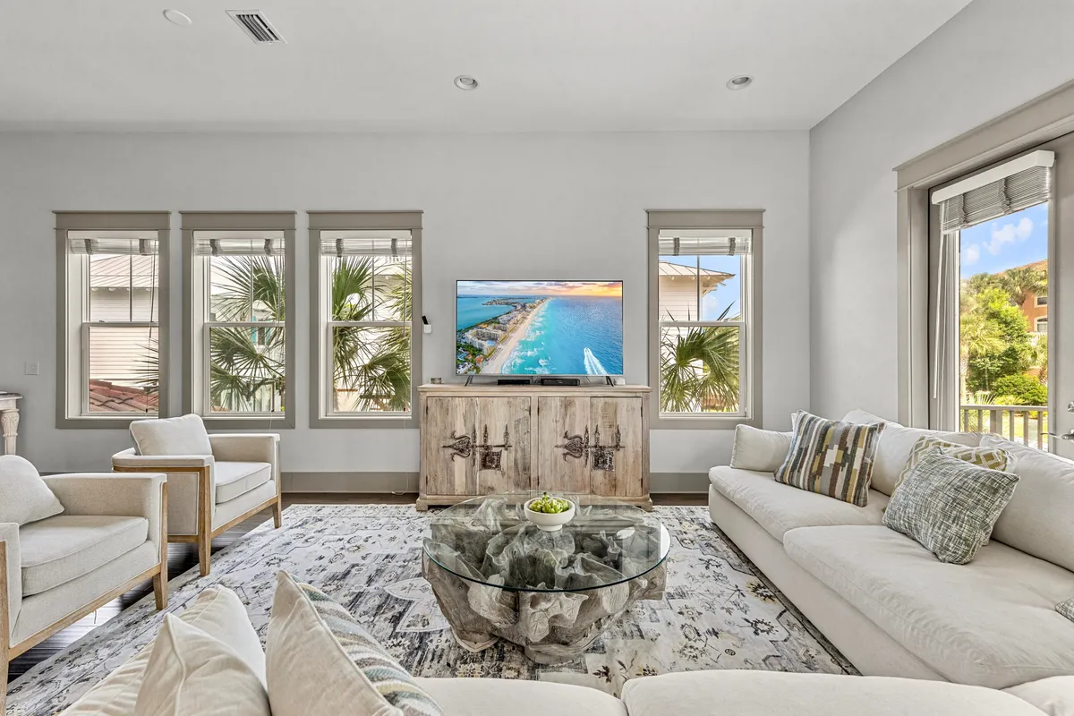 A spacious, inviting living room with large windows framing palm tree views. The area features a comfortable sofa and armchairs, a large-screen TV showing a beach scene, and a glass coffee table with decorative accents. The overall mood is casual and comfortable, perfect for both relaxation and group gatherings.