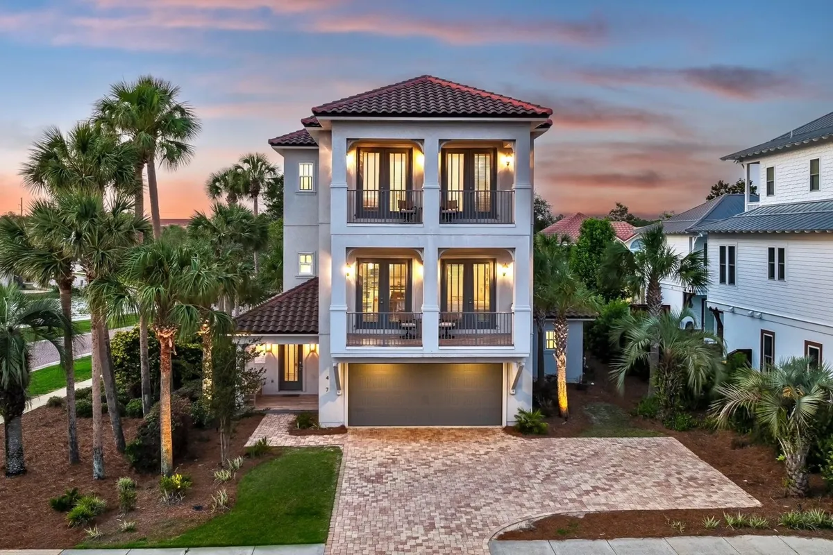 The image showcases a grand, three-story luxury home with multiple balconies glowing warmly in the evening light. Palm trees and lush landscaping frame the brick driveway, creating an inviting tropical entrance. The home's exterior radiates elegance and comfort, with spacious outdoor spaces perfect for relaxing or socializing in a serene, gated community.