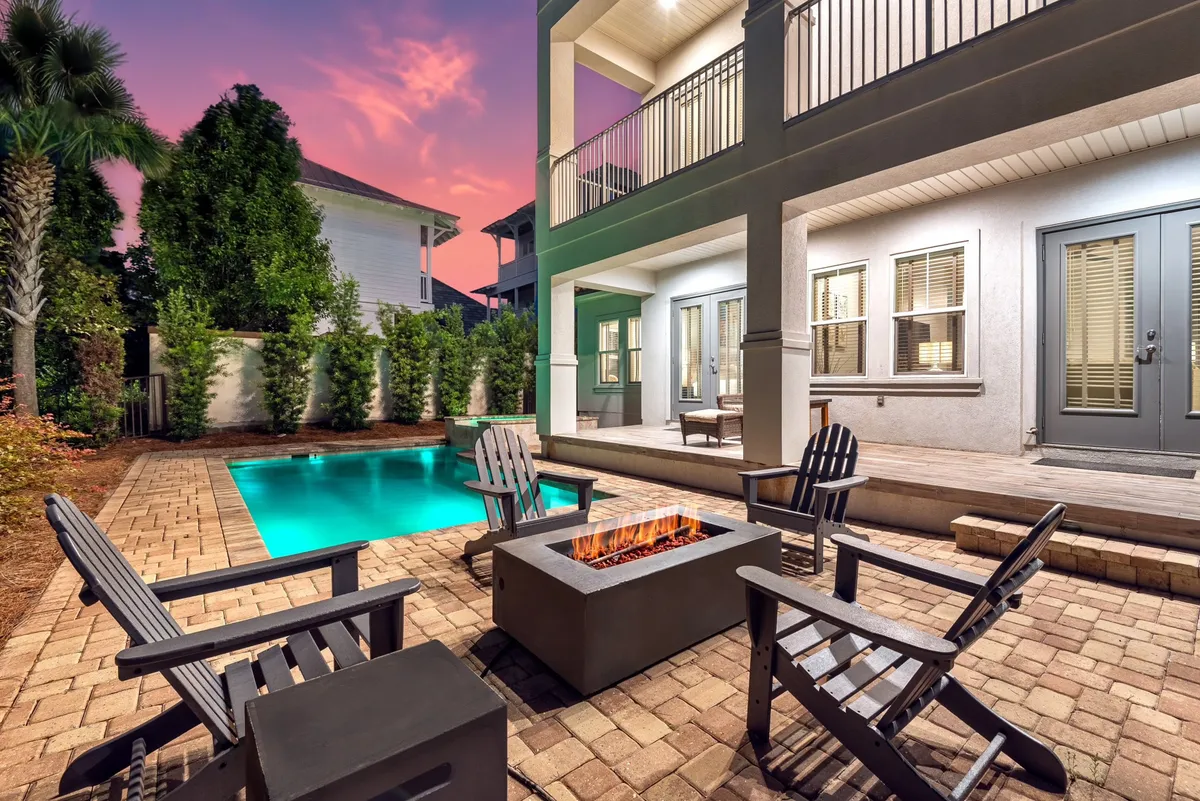 This image features a private heated pool illuminated for evening enjoyment, surrounded by lush greenery and palm trees. A fire pit encircled by Adirondack chairs is prominently positioned beside the pool, with a covered patio seating area completing the scene. The ambiance is tranquil and inviting, perfect for both lively gatherings and quiet reflection.