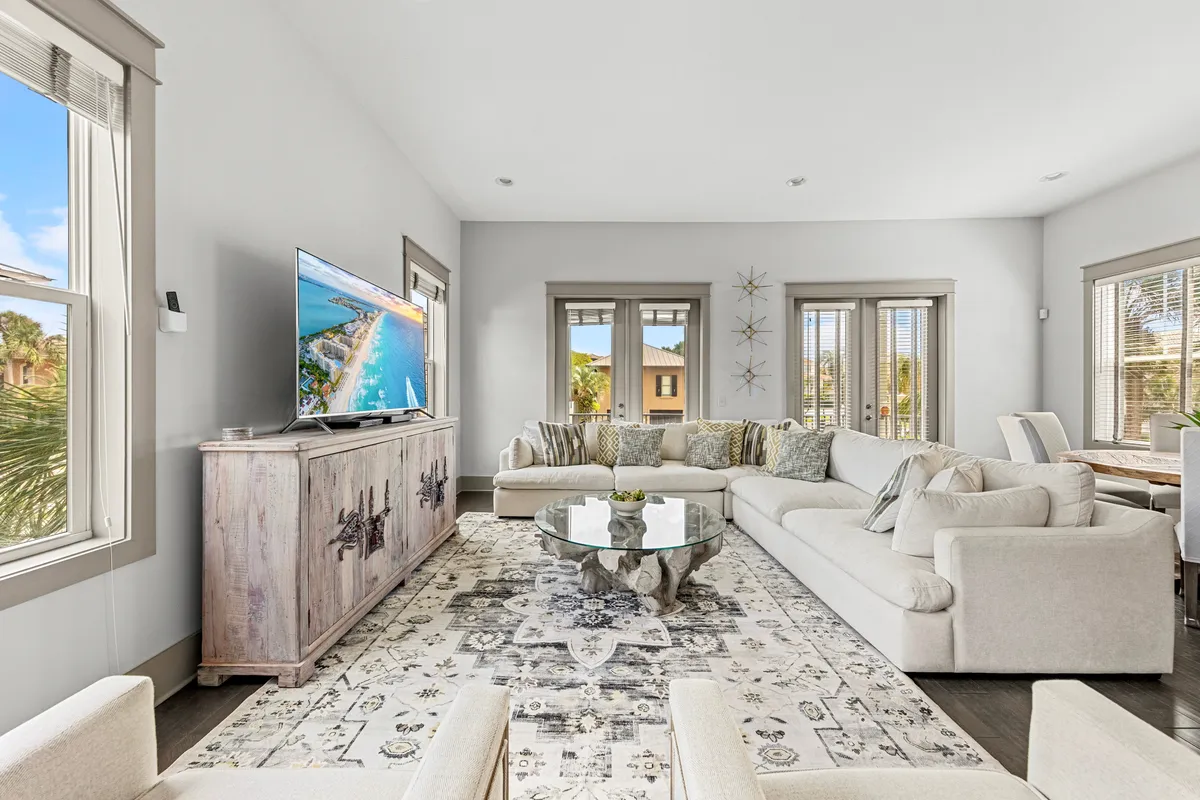 A cozy, sun-filled living room with a large sectional sofa, a unique driftwood-style glass coffee table, a flat-screen TV on a rustic console, and a dining area visible in the background. Multiple windows provide abundant natural light and views of the outdoors.