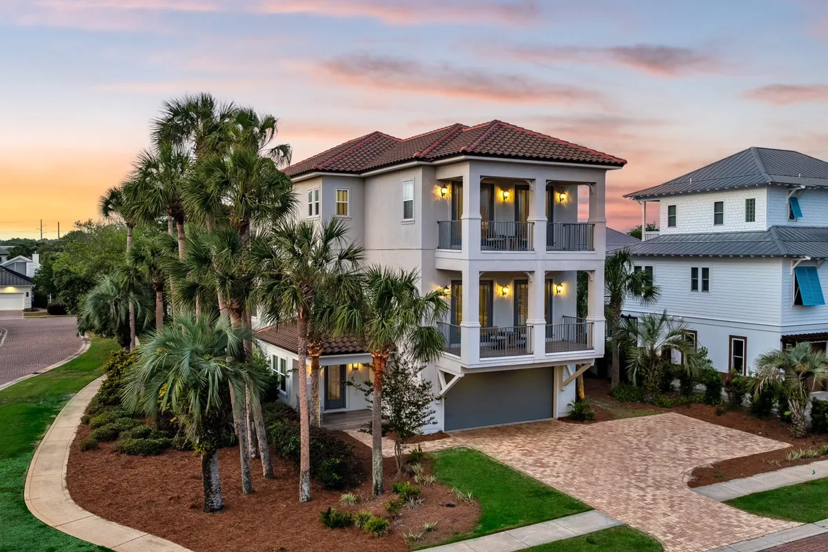 A three-story luxury home surrounded by tall palm trees, with balconies on each floor and warm outdoor lighting illuminating the entrances. The well-maintained driveway and landscaping add to the upscale, welcoming mood, hinting at both comfort and proximity to outdoor adventures.