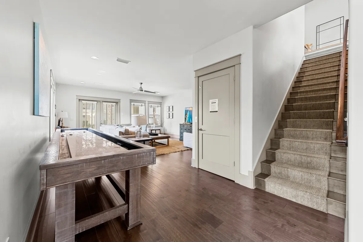 A welcoming, open-plan space featuring a shuffleboard table, a bright living area with sofas and large windows, and a staircase leading upstairs. The room is filled with natural light, inviting both play and relaxation.
