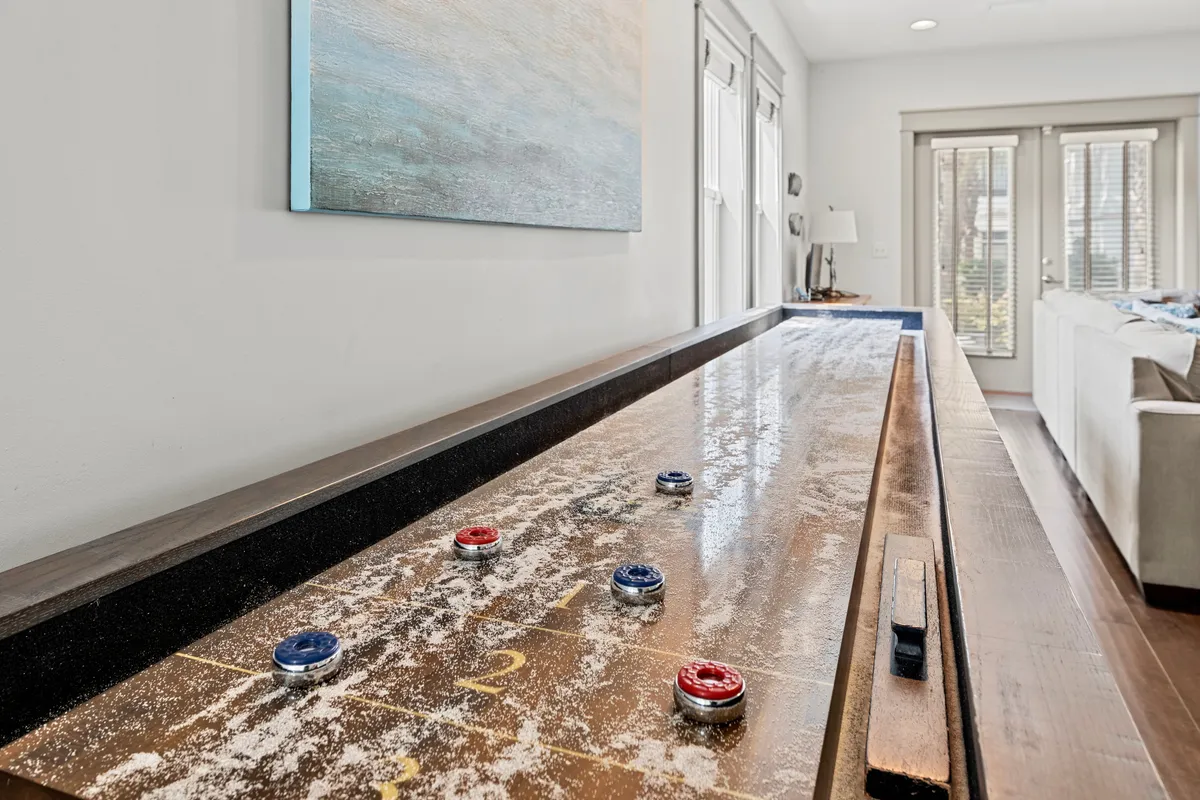 A bright, airy indoor recreation area showcasing a shuffleboard table with game pieces, comfortable seating in the background, and large windows that fill the room with natural light. Modern artwork adds a touch of style to the space.