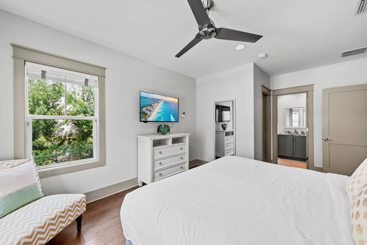 A peaceful king bedroom with a large bed dressed in crisp white linens, a window with garden views, an armchair with pastel pillows, and a wall-mounted flat screen TV. Natural light creates a restful and airy atmosphere.