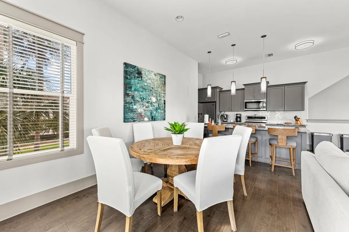 A round wooden dining table surrounded by white upholstered chairs sits next to a modern kitchen with gray cabinetry and a large window showing palm trees. The setting is intimate and welcoming, ideal for shared meals and special occasions.
