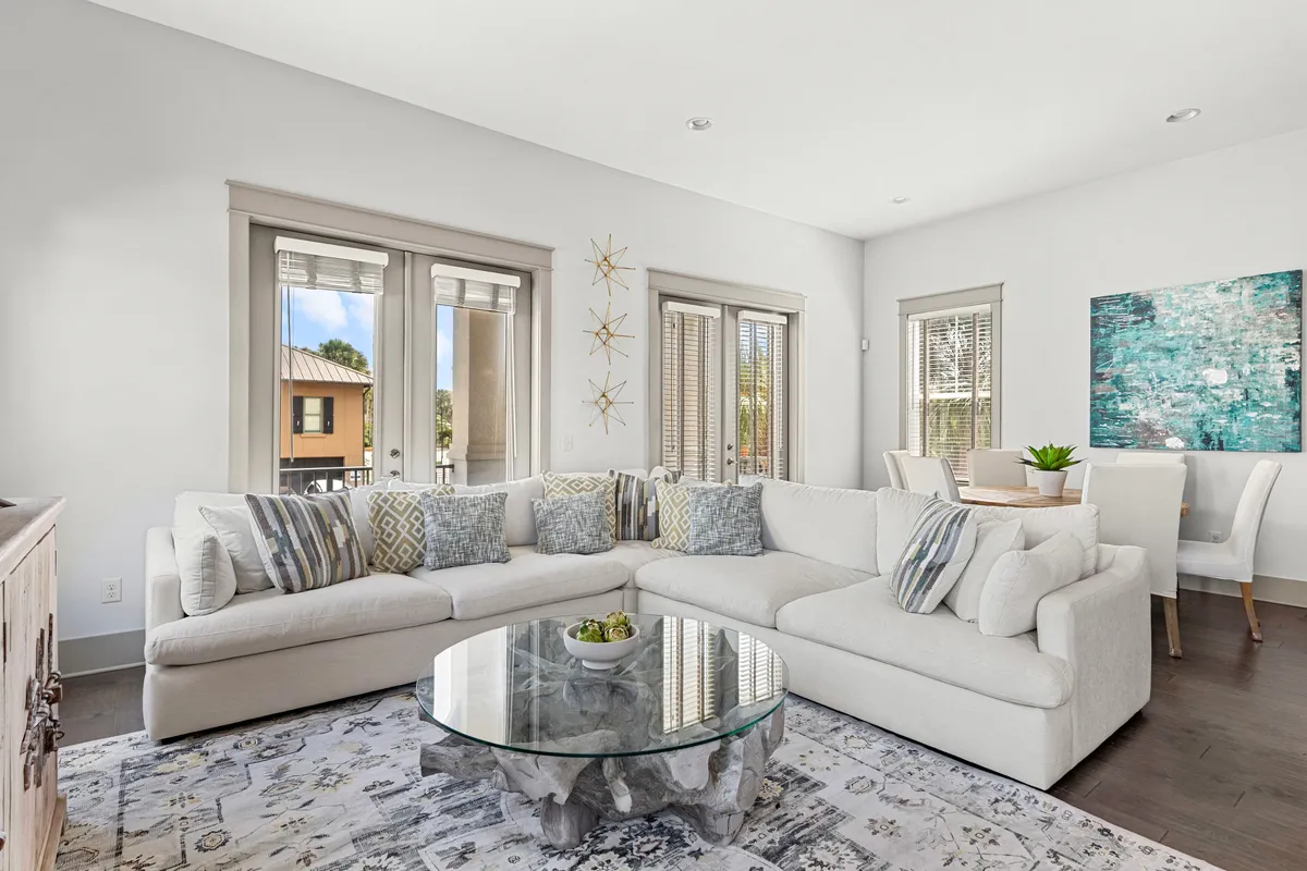 A bright, airy living room with a large white sectional sofa, round glass coffee table with a driftwood base, and a dining area with white chairs and a wooden table. Decorative wall art and starfish accents add a coastal touch, while windows invite in natural light and views of the outdoors.