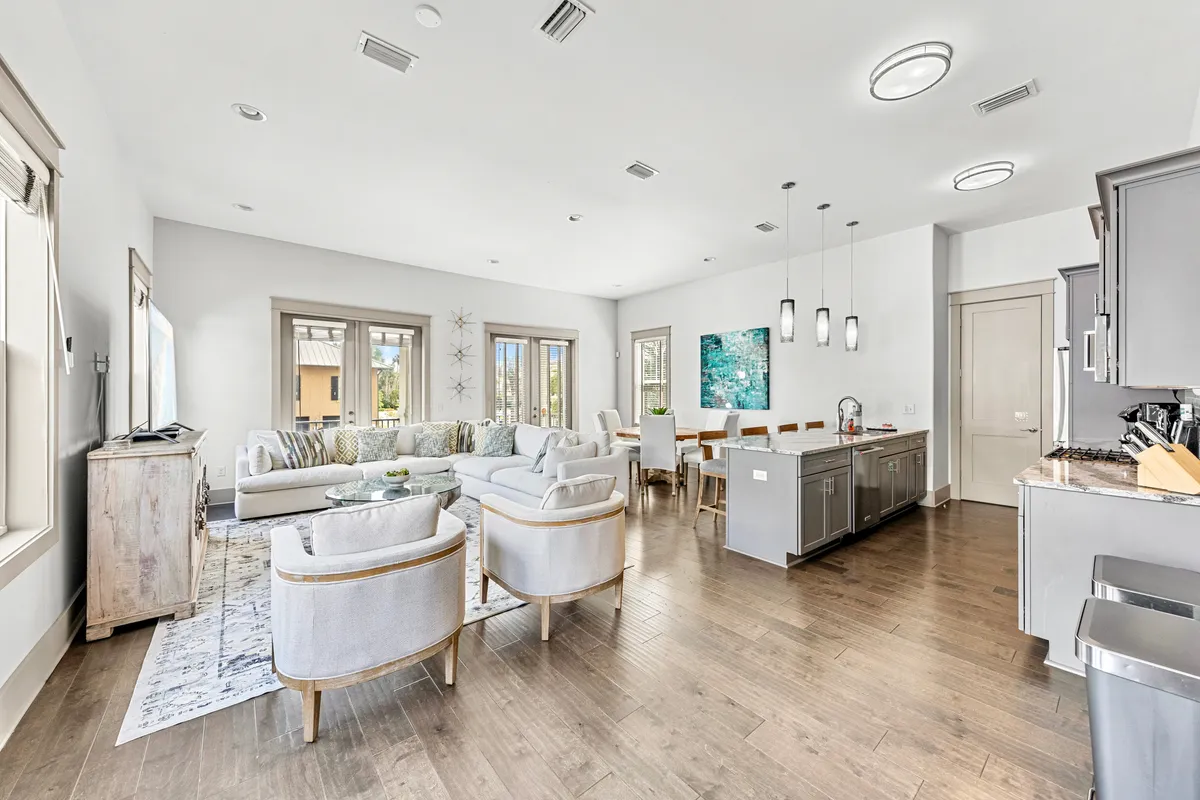 This image features a bright, open-concept living space with a large sectional sofa, a kitchen island with pendant lighting, a dining area with artwork, and a flat-screen TV on a rustic stand. The living room and kitchen flow seamlessly together, with plenty of room for socializing and group activities.