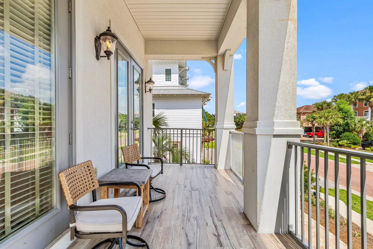 This exterior image features two cushioned wicker chairs and a wooden table on a balcony, framed by white railings and palm trees. The view overlooks neighboring houses within a quiet, gated community, conveying a sense of peaceful exclusivity.