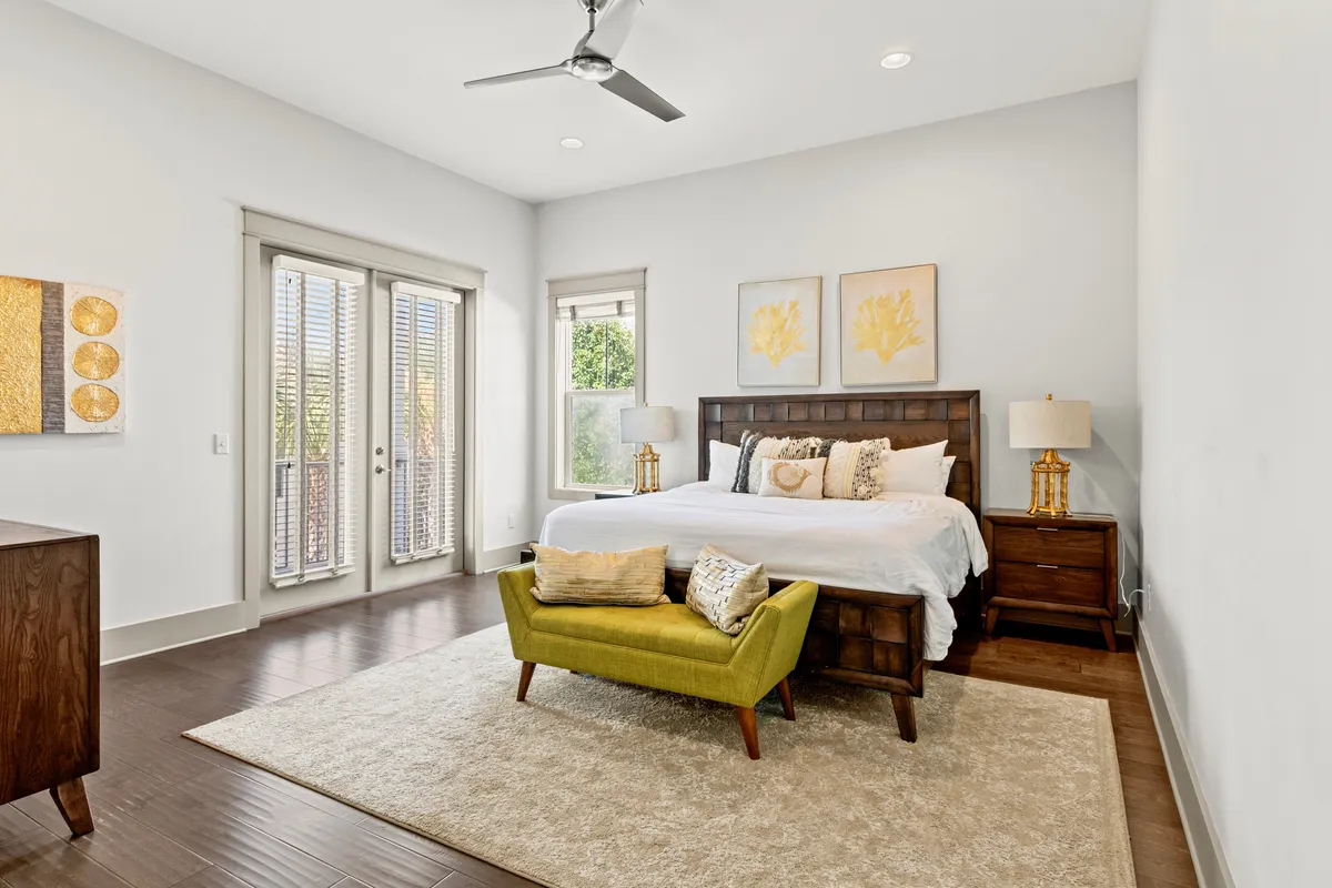 A tranquil king suite featuring a king-size bed dressed in white linens and decorative pillows, complemented by an olive green upholstered bench at the foot. French doors with blinds open onto a private balcony, and golden coral-themed wall art hangs above the bed, all bathed in gentle natural light.