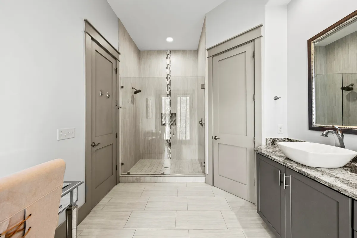 A modern, spa-inspired bathroom featuring a glass-enclosed shower with decorative tile strip, a vessel sink atop a granite countertop, a large framed mirror, and neutral cabinetry. The space feels calm and refreshing, perfect for personal care and relaxation.
