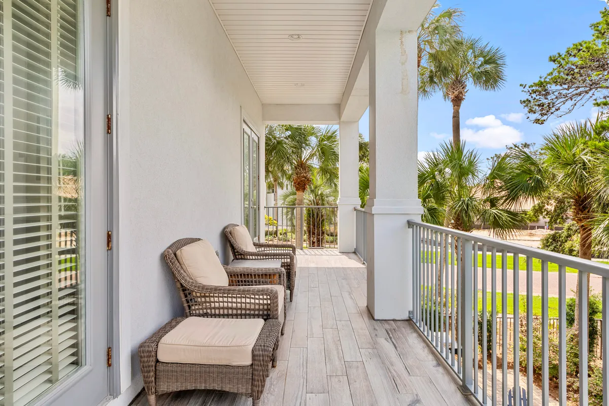 A spacious balcony with wicker lounge chairs overlooks palm trees and a bright blue sky. The tiled floor, white railings, and columns evoke a serene, tropical retreat ideal for unwinding after a day of complimentary activities.