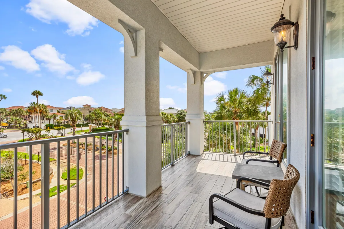 A tranquil exterior scene featuring a covered balcony with comfortable seating, overlooking palm trees, a gated community, a paved walkway, and a landscaped garden. The view extends to quiet streets and distant buildings, evoking a sense of peaceful outdoor living.