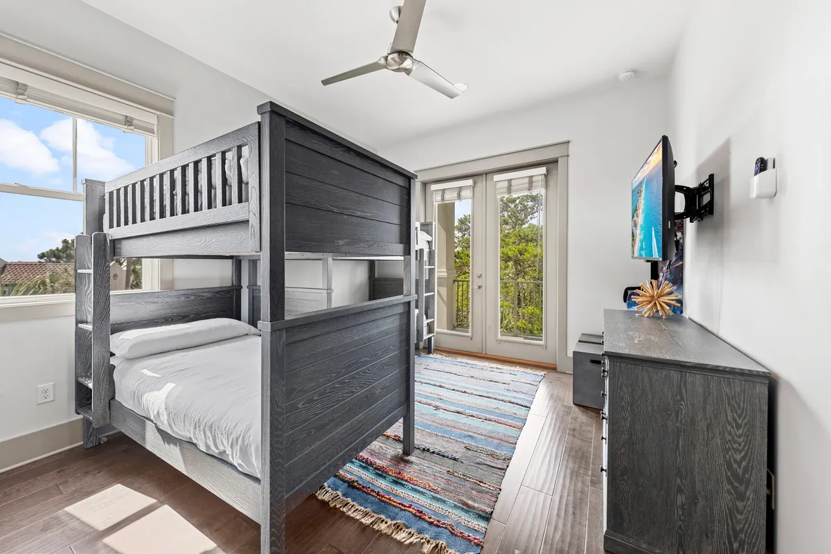 A bright and playful bunk room with black wooden bunk beds, a colorful striped area rug, French doors leading to a balcony, and a wall-mounted flat screen TV. Abundant natural light floods the space, making it inviting for children or the young at heart.