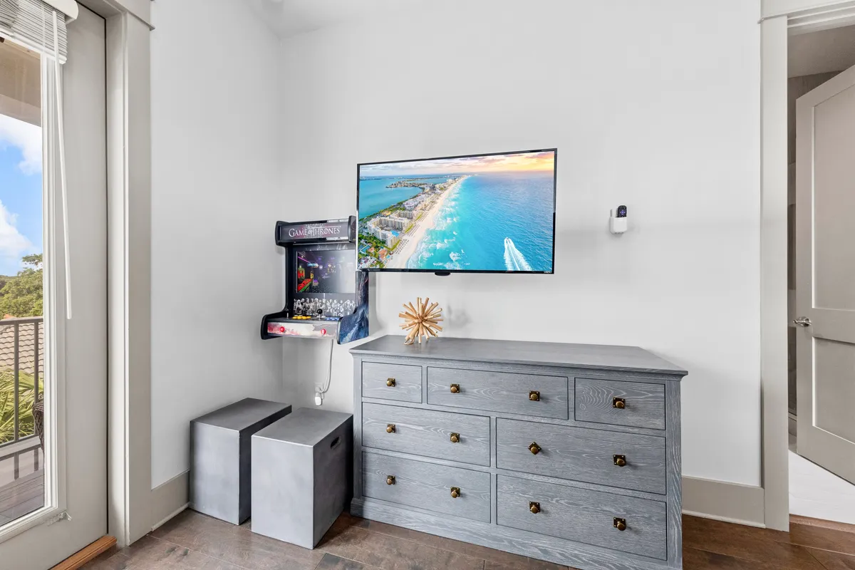A cozy and modern entertainment room featuring a wall-mounted flat screen TV displaying a beach aerial view, a game machine mounted on the wall, a grey wooden dresser for storage, and two cube seats for casual relaxation or play. The space exudes a relaxed, playful ambiance ideal for family fun.