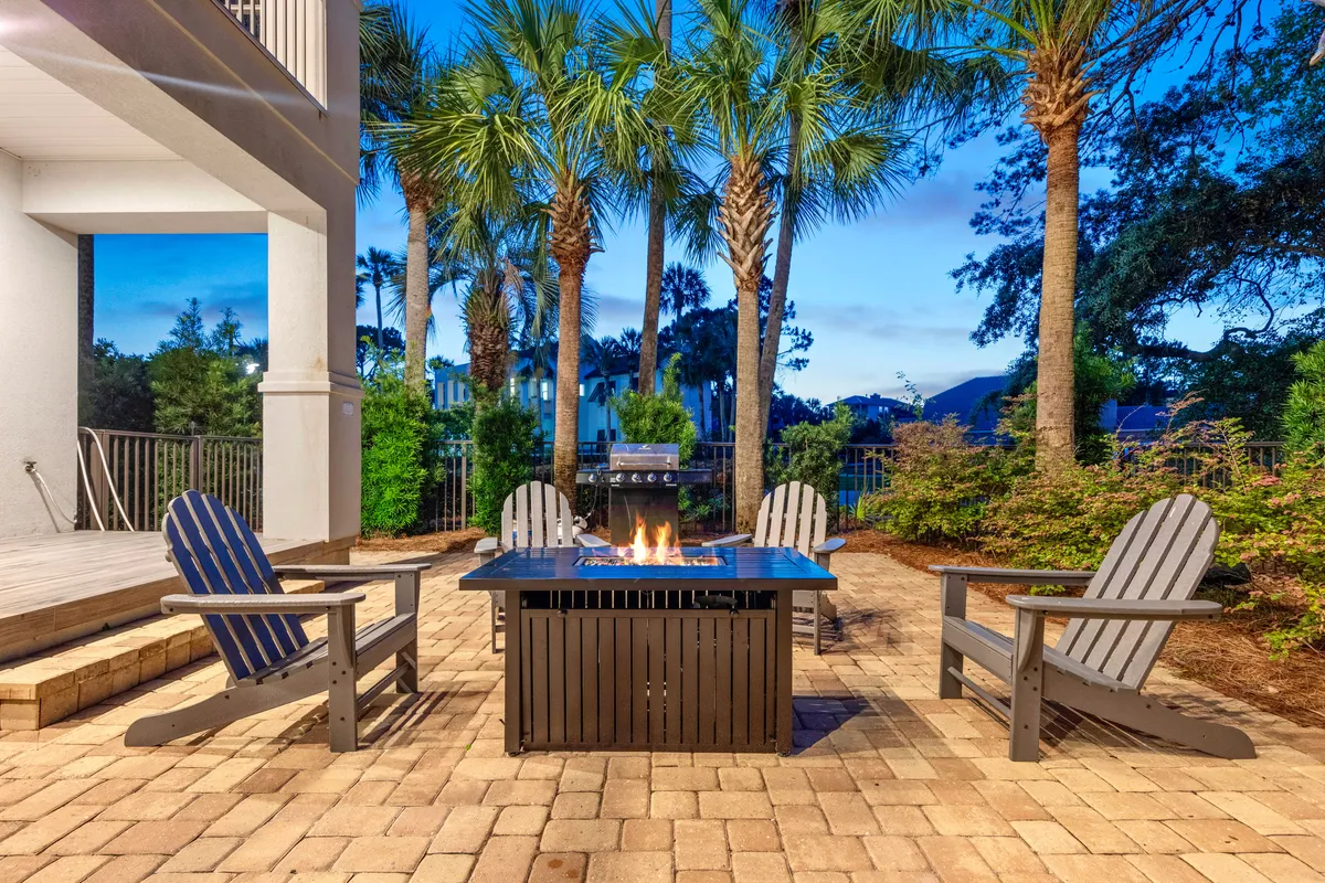 This image spotlights a central fire pit surrounded by four Adirondack chairs, with a grill in the background and tall palm trees enclosing the patio. The area is clearly designed for outdoor gatherings, relaxation, and cooking in a lush, private setting.
