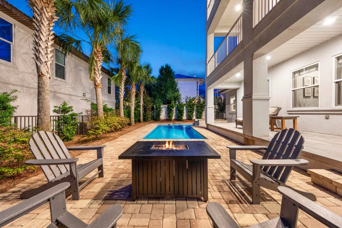 A private heated pool extends alongside a patio, where a glowing fire pit is surrounded by Adirondack chairs under tall palm trees. The ambiance is cozy and inviting, perfect for evening relaxation or intimate group gatherings in a tropical, upscale backyard.
