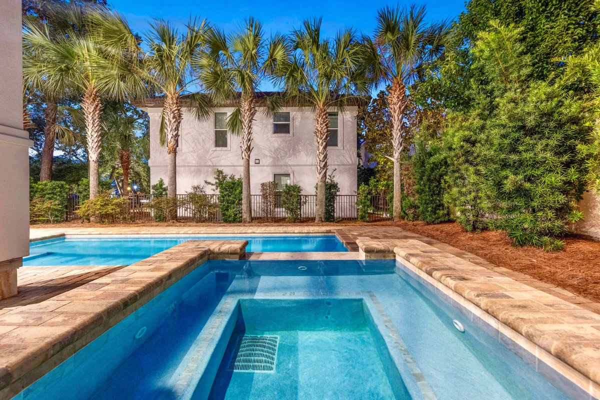 A serene daytime scene centered on a private heated pool and adjacent hot tub, bordered by tall palm trees and stone decking. The tranquil setting offers ample space for swimming, relaxing in the jacuzzi, and socializing in a tropical environment.