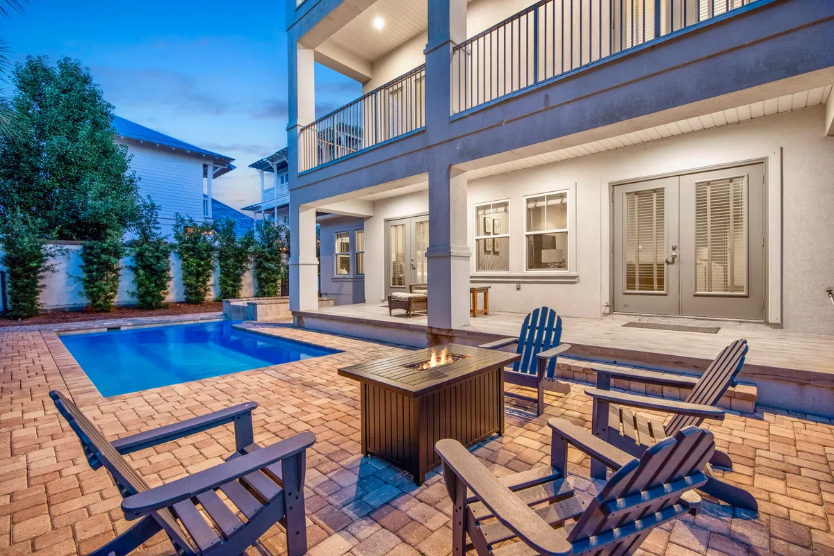 This exterior image features a private heated pool with crystal-clear blue water, an inviting fire pit surrounded by Adirondack chairs, a covered patio area with ample seating, and a two-story balcony overlooking the pool area. The setting is lush with palm trees and landscaping, creating a tranquil yet lively atmosphere.
