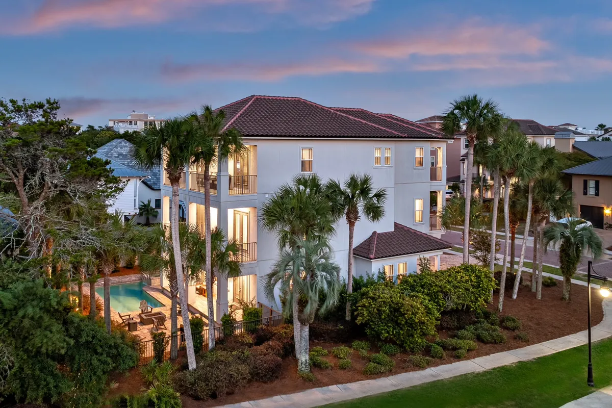 An exterior evening view of a three-story luxury home with a Mediterranean-style tiled roof, palm trees, a private heated pool area, and softly glowing indoor lights. Outdoor dining and lounge furniture are visible, hinting at both relaxation and social gatherings.
