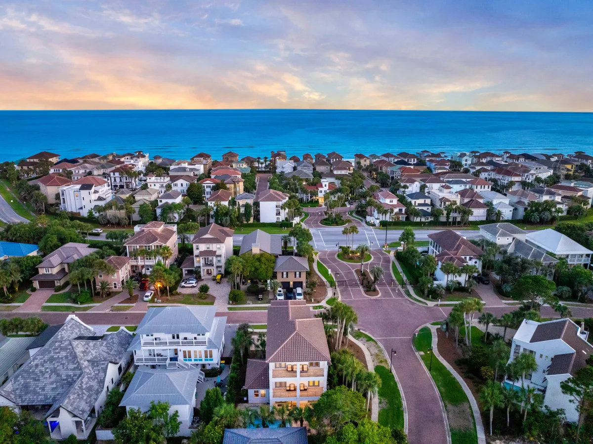 Rows of upscale houses with lush landscaping and palm trees line calm, winding residential streets, with the blue ocean horizon visible in the background. The scene conveys the exclusivity and tranquility of the Destiny East community, hinting at the range of activities and amenities available, from strolls to beach outings.