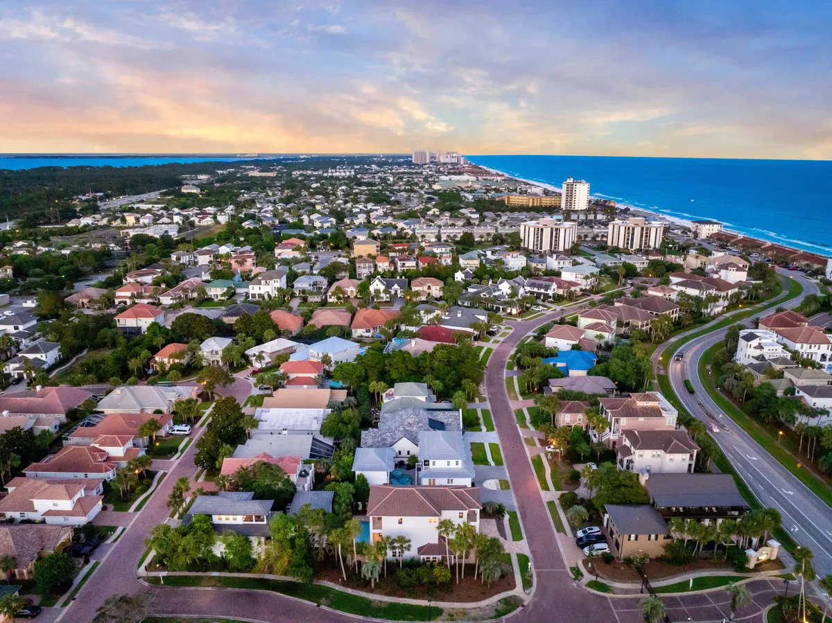 A sweeping exterior view of a tranquil coastal neighborhood with a curved road leading through rows of upscale houses. The blue ocean coastline stretches to the horizon under a sunset sky, creating a warm, inviting atmosphere. The image highlights proximity to the beach, walkability, and the relaxed vibe of the area.